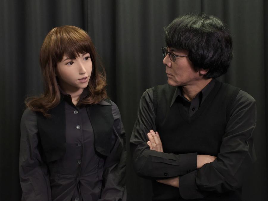 Hiroshi Ishiguro, a man with black hair, glasses and a black outfit looks at Erica, a female humanoid robot with golden skin, almond shaped eyes, and shoulder length brown hair.