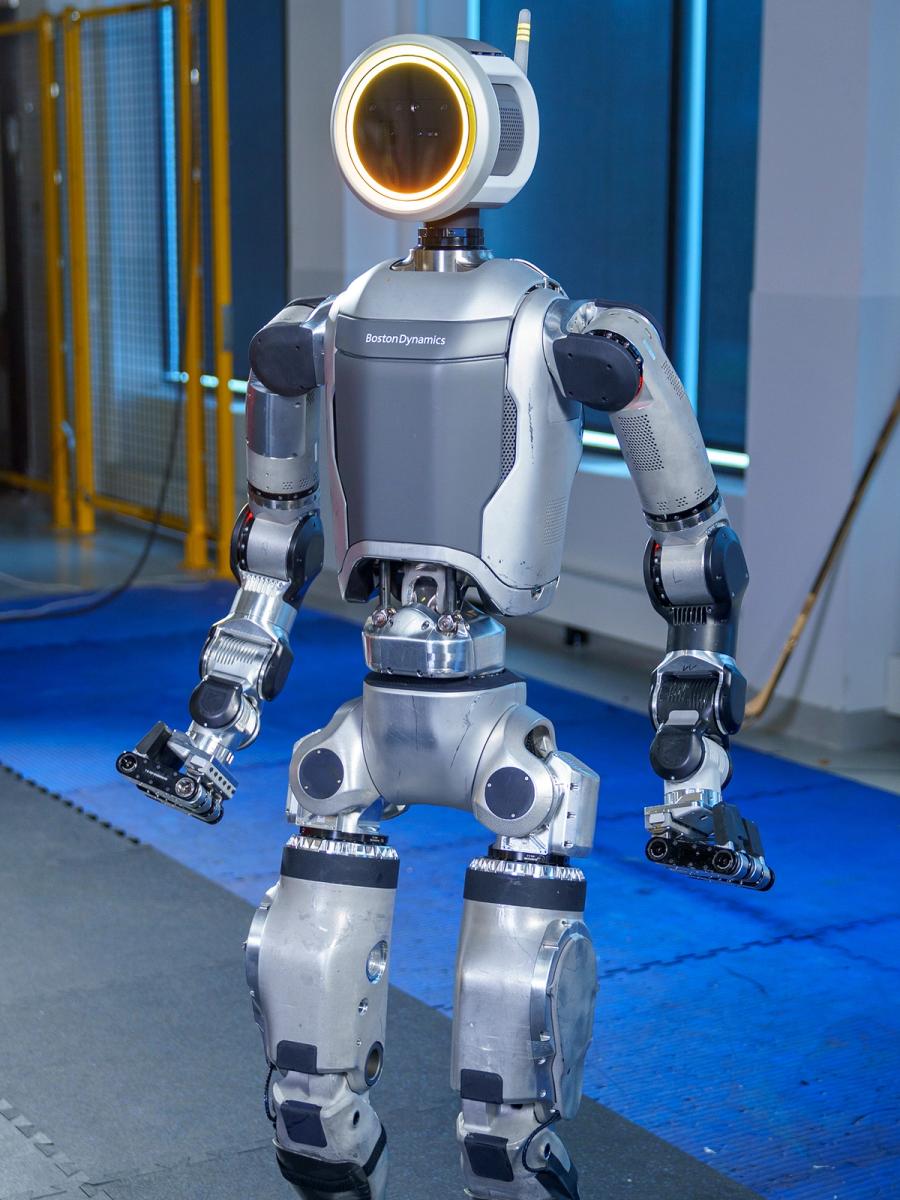 Three quarter view of Atlas, a grey and silver bipedal robot with a circular head that glows yellow, and long arms and gripper hands.