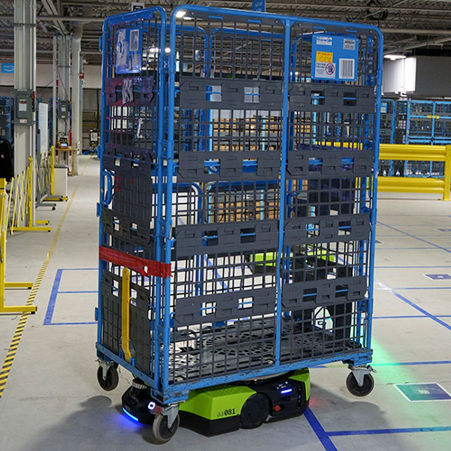 A squat green mobile robot carries a large shelved unit in a factory.
