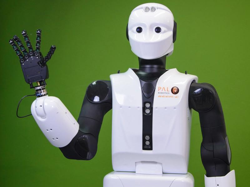 A shiny white robot with a simple face involving two eyes, and two sensors in its forehead. It's black and white arm ends in a five digit hand, which the robot holds up.