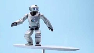 Knee-high humanoid robot Qrio, which has a silver body with round eyes, balances on a narrow board as if it were surfing.
