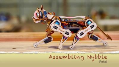A small cat robot made of laser-cut wood and electronics walks on a wooden table with the words Assembling Nybble shown on bottom.