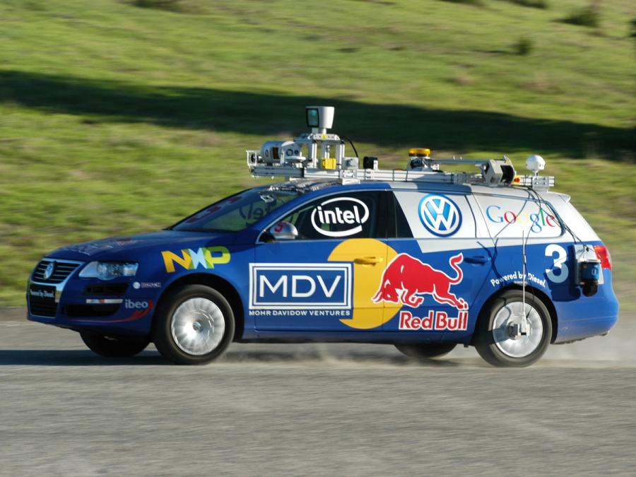 A blue, logo covered self-driving car with a roof rack full of electronics drives on asphalt.