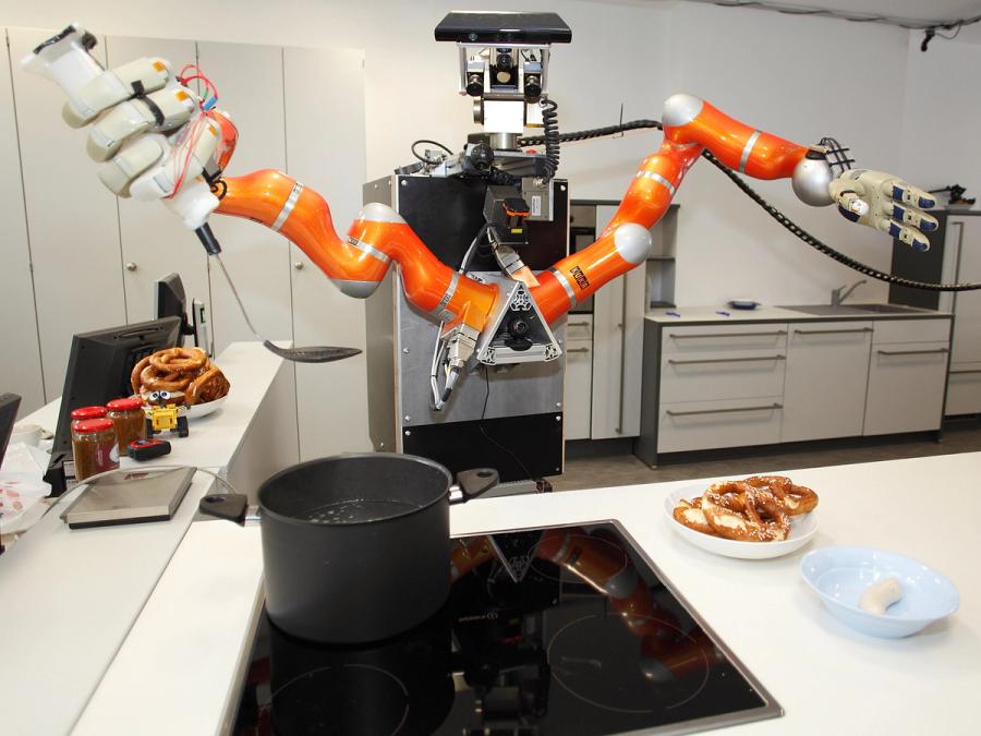 The robot stands in the kitchen over a hot pot. One hand holds a ladle. There is a plate of food on the counter.