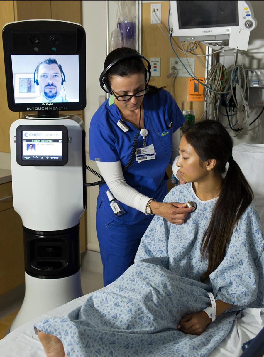 A telepresence robot with a large display on top which shows a doctor's face next to a nurse working with a bed-bound hospital patient.
