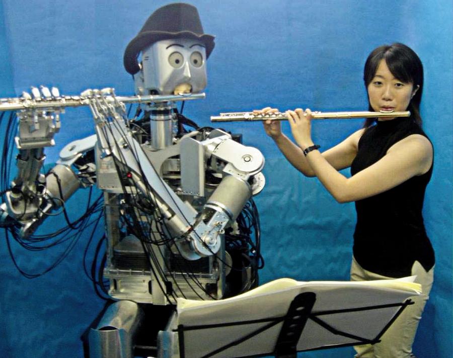A human sized humanoid robot and a human female each play a flute while sitting side-by-side looking at sheet music on a stand.