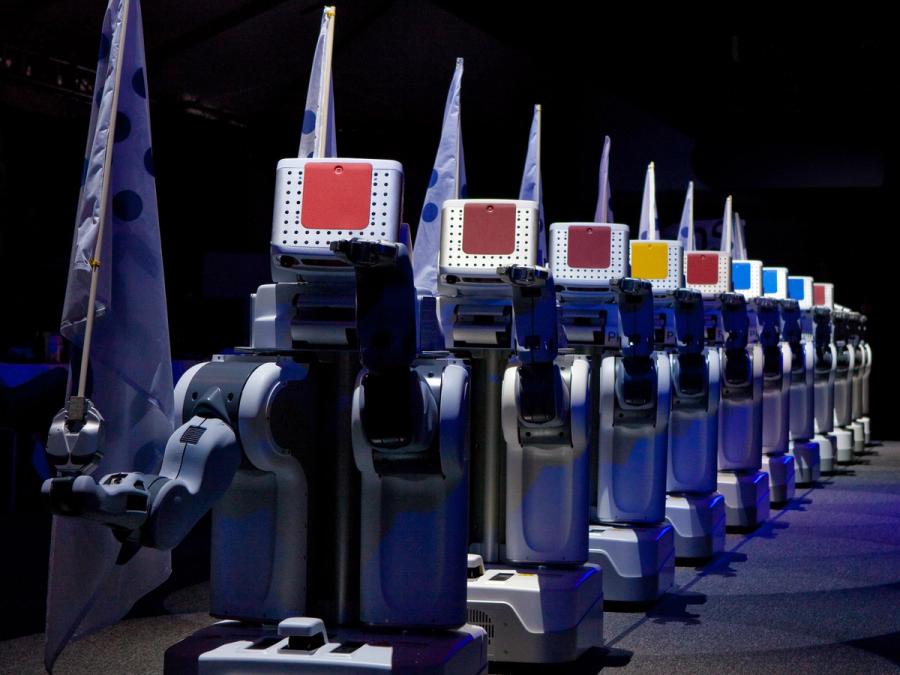 A row of a dozen PR2s. Each holds a white box with a colored square on top, and carries a flag in one hand. The other hand on each robot is raised in unison.