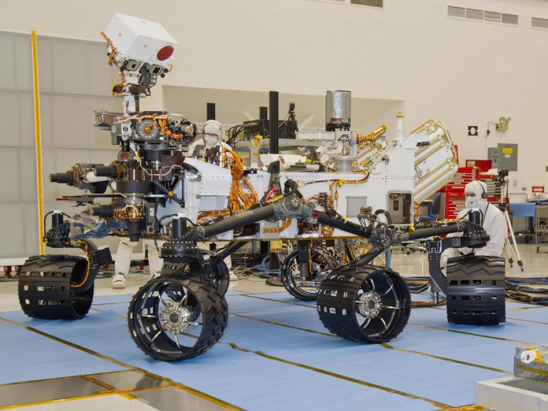 A person in a clean room bunny suit kneels next to a large, six-wheeled robotic vehicle equipped with cameras and instruments. 