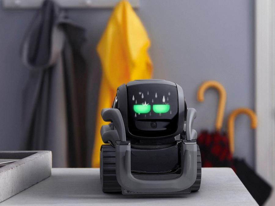 A simple, compact, black wheeled robot smaller then the palm of a hand with two glowing green eyes sits on surface in a home.
