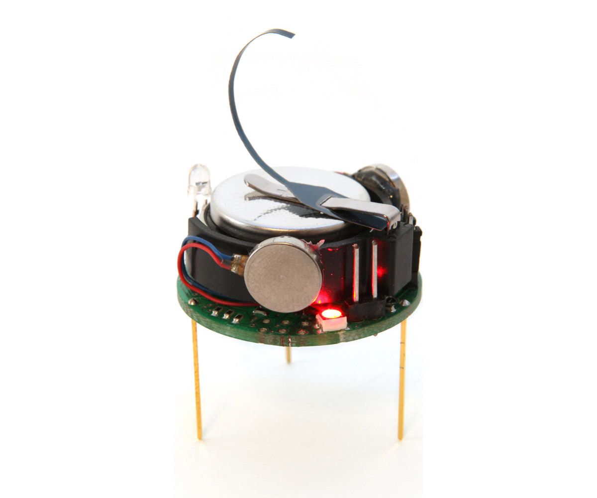 A spinning view of a single 5.5 cm tall kilobot which has 3 toothpick like yellow legs, electronics on a round circuit board and a curved metal piece sticking out of the top.