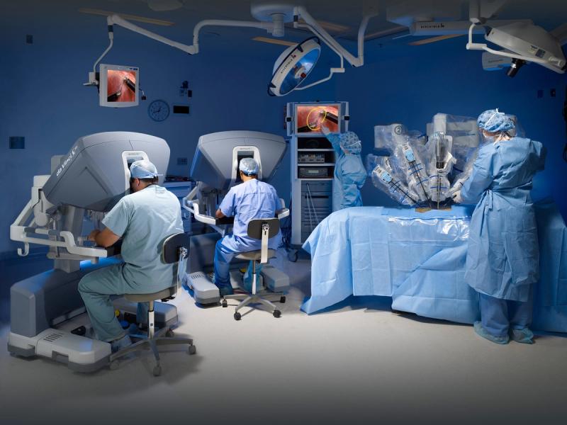 The Da Vinci robotic surgical system used in an operating room, with its multiple robotic arms in position over a patient, while surgeons monitor the procedure using 3D visualization displays.