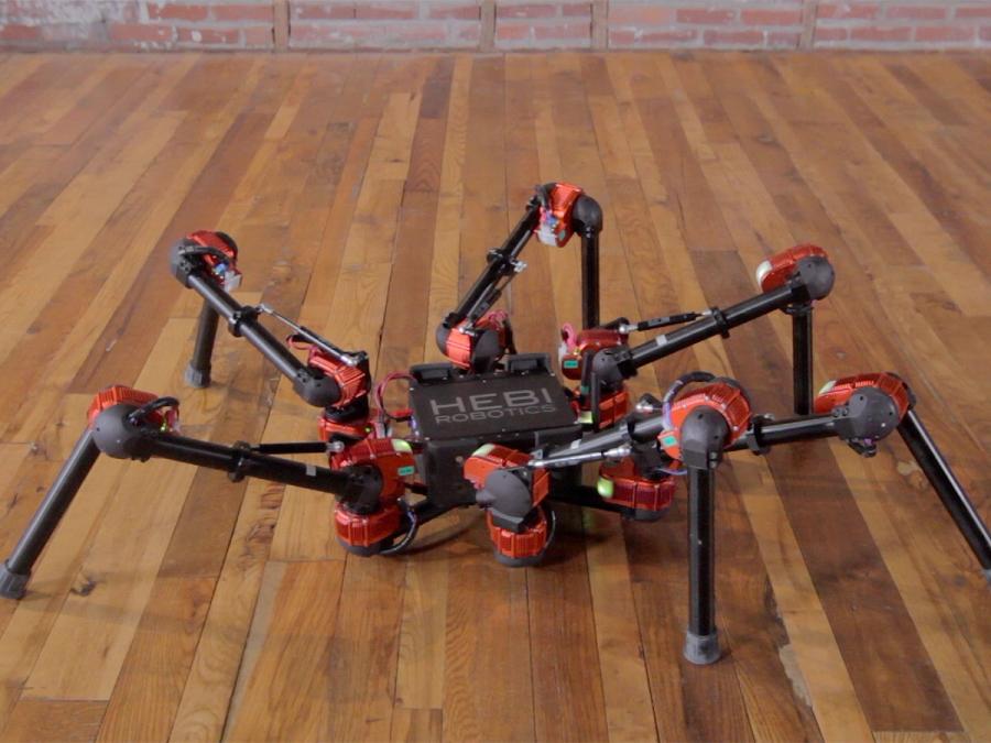 A hexapod robot is seen with it's center unit lowered to the wood floor, while it's legs remain bent.