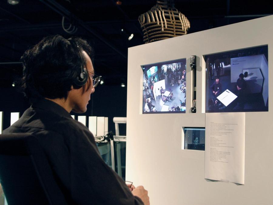 A man with headphones on looks at several video screens on a wall in front of him.