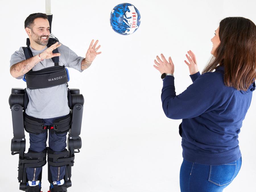 A smiling man in a tethered exoskeleton holds his hands out to catch a ball being thrown by a woman wearing a blue shirt and jeans.