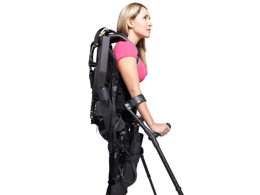 A blonde woman in a pink shirt wears a black exoskeleton backpack suit, and is supported by crutches attached to her arms.