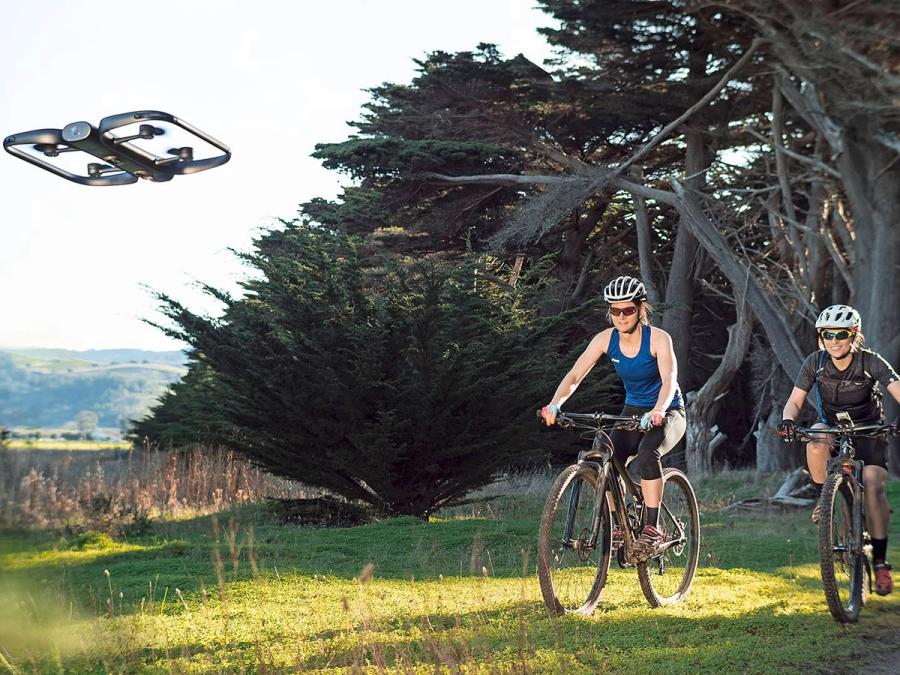 Two helmeted people bike in a scenic landscape while a black drone flies in front of them.