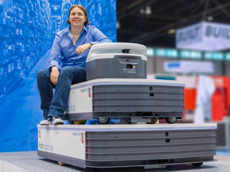 A woman in blue shirt and jeans sits on middle level of three stacked robotic platforms of varying sizes. 