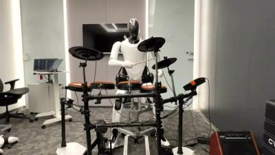 A humanoid robot with white and black body holds drumsticks while sitting on an electronic drum kit.
