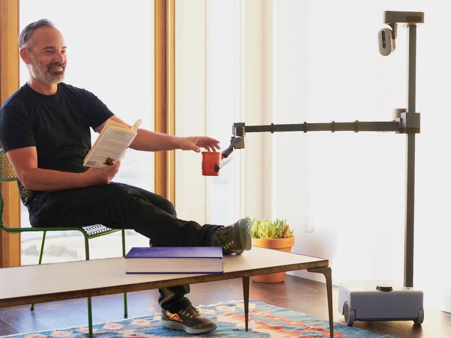A mobile robot extends its telescoping arm to deliver a mug to a man who is sitting and reading a book.