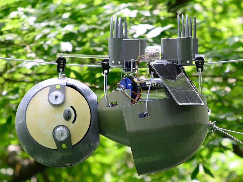 The SlothBot robotic sloth hangs upside down from a wire amid green leafy trees outside. It has a solar panel and other electronics.
