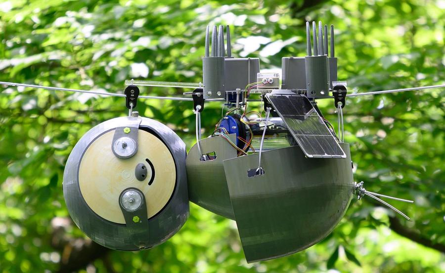 A robotic sloth hangs upside down from a wire amid green leafy trees outside. It has a solar panel and other electronics.