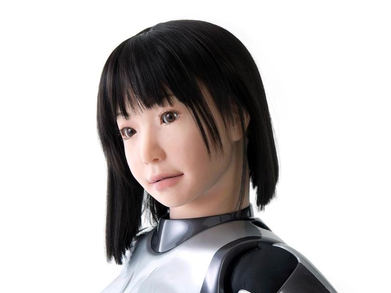 Close-up of a female robot with a realistic appearance including peach skin, pink lips, brown eyes and a black bob hairdo.