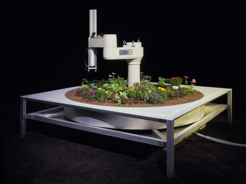 A white robotic arm with a vertical appendage sticks out from a flower filled circular garden in a platform like a silver coffee table.