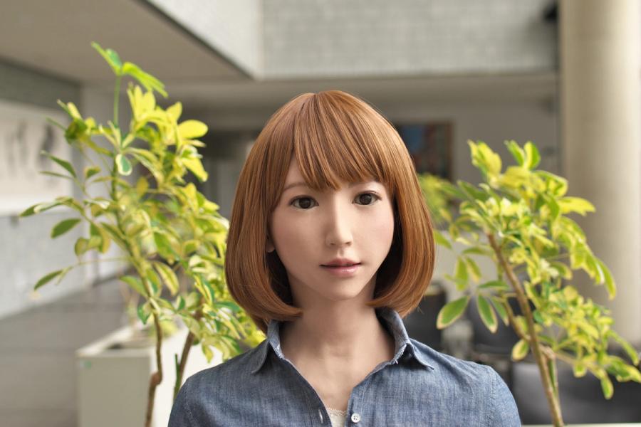 Female robot Erica wearing a new long bob hairstyle that is lighter brown while sitting in a brightly lit atrium with plants in the background.