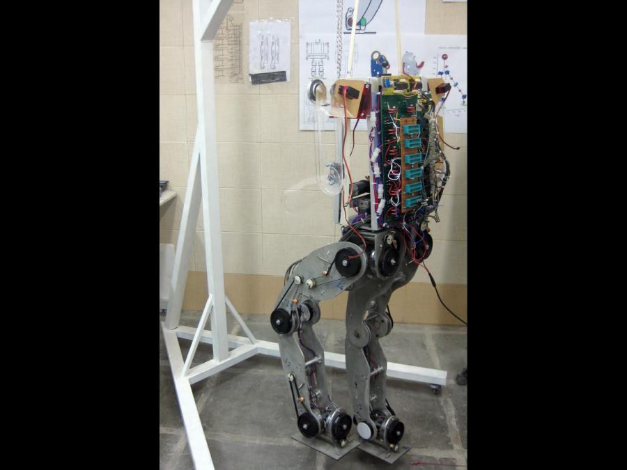 A prototype humanoid consisting of a metal frame, electronics, and wires takes a step.