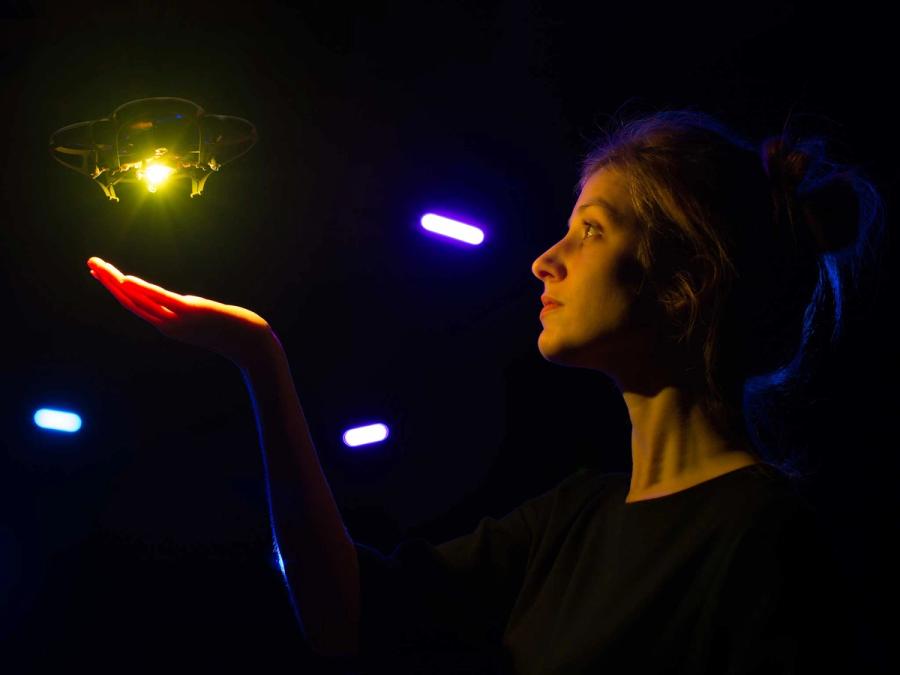A woman in a dark room holds out her palm. A small drone with a yellow light on the bottom hovers above her hand.
