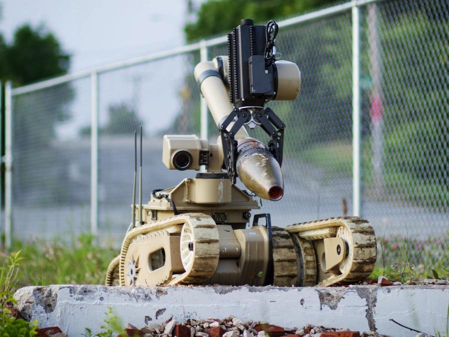 A beige wheeled robot with tank-like treads with a bent articulated arm featuring a strong black gripper hand rolls over a sidewalk.