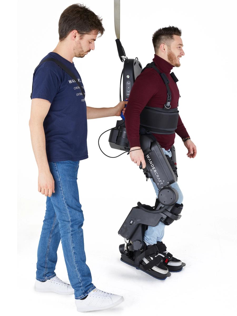 A smiling man takes a step wearing in a Wandercraft exoskeleton that includes strapping around his legs and torso. A second person holds a strap on his side.