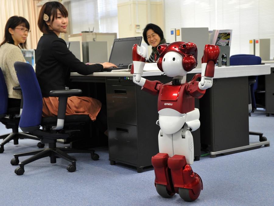 Red and white child sized robot rolls through an office, while three office workers look at it.