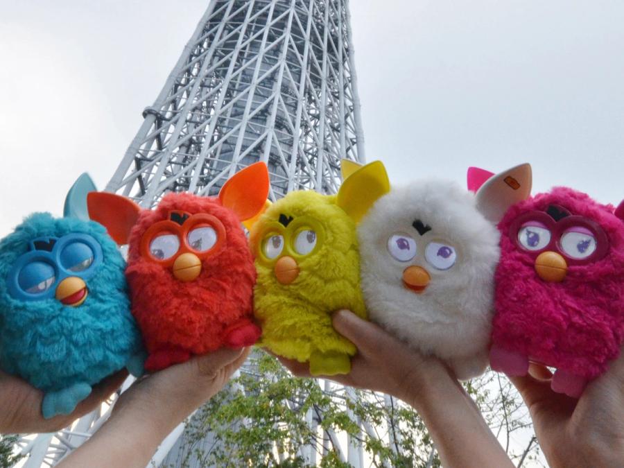 Two sets of hands hold up five Furbys, furry owl-like toys in blue, red, yellow, white and pink.