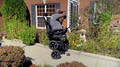A man, sitting on an iBOT powered wheelchair balancing on two wheels, takes a photograph of a dog on the front lawn of a house.