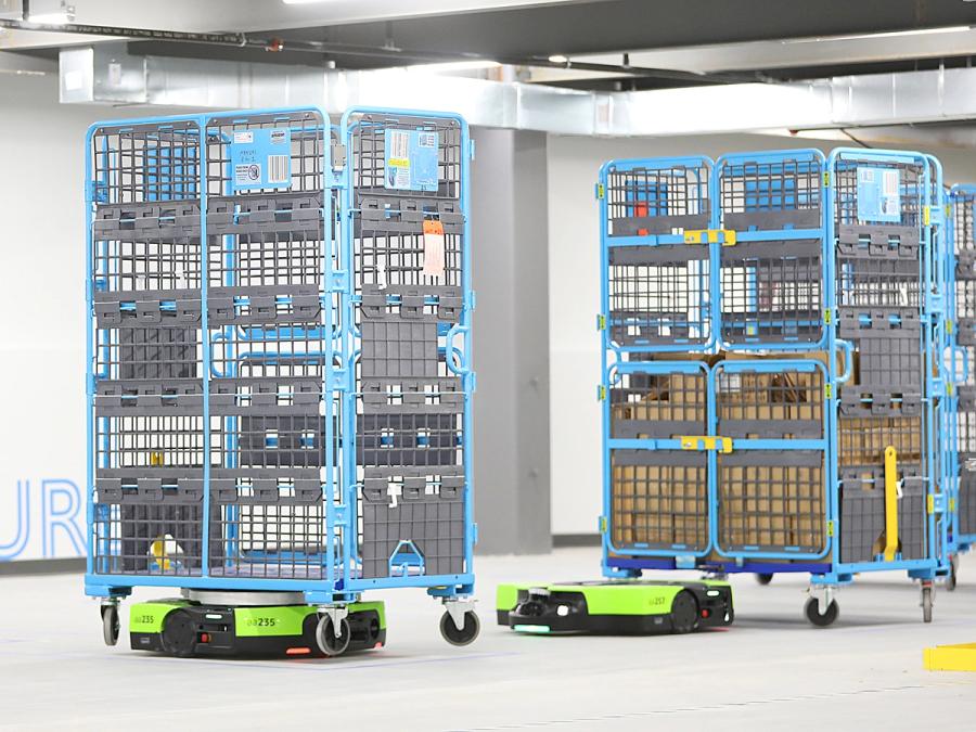 Two low to the ground green mobile robots carry blue multi-level shelving units through a warehouse.