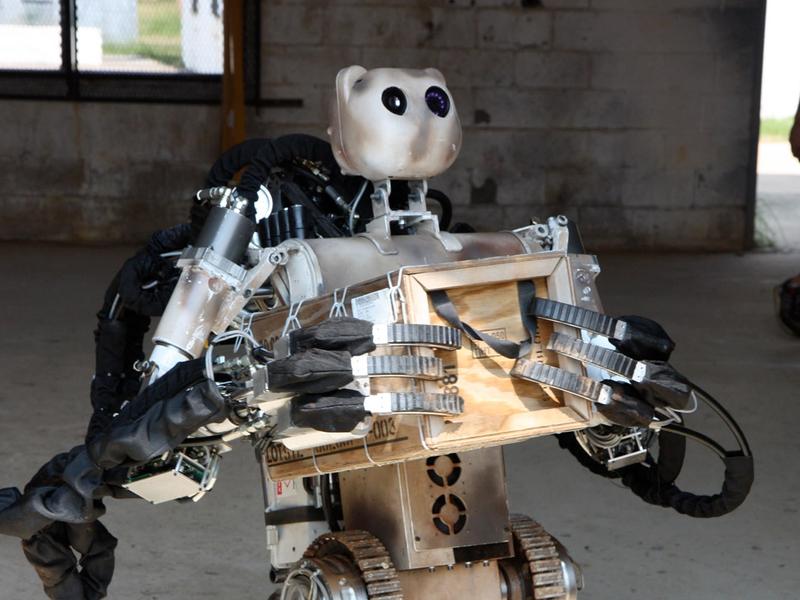 Bear, a wheeled, two handed, three fingered robot, with a bear shaped head holds a wood crate to its torso.