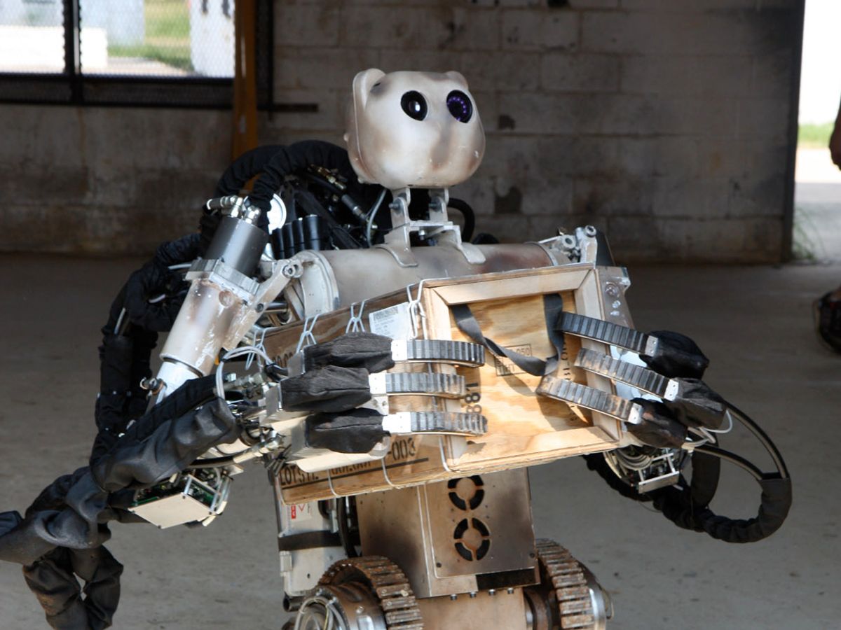 A wheeled, two handed, three fingered robot, with a bear shaped head holds a wood crate to its torso.