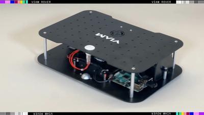 A squat robot made of black metal rectangular sheets with electronics, sensors, and two wheels, drives on a smooth white surface.