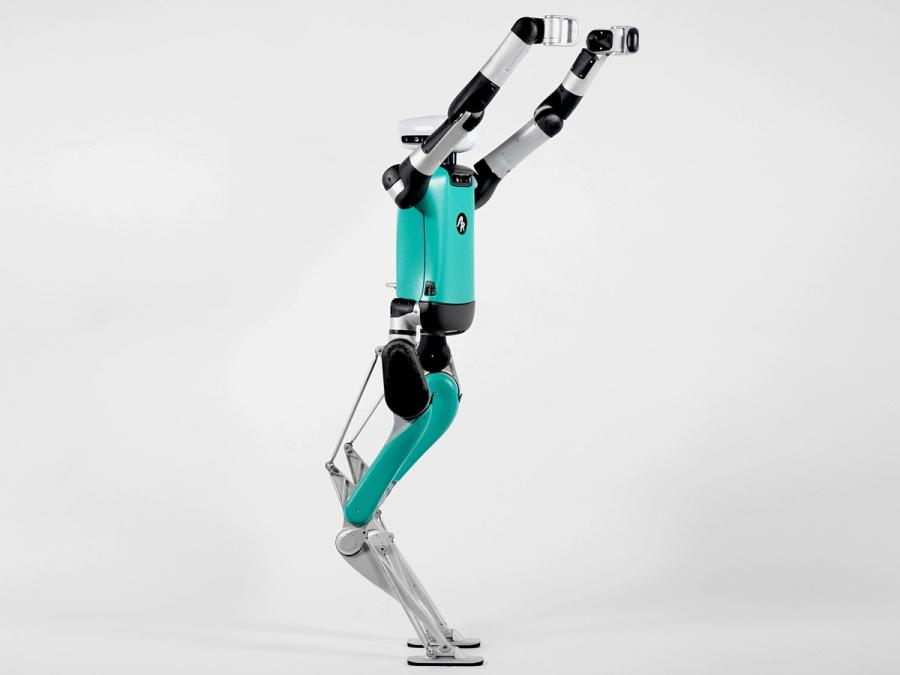 A full length view of Digit shows a teal, silver and black bipedal robot with jointed legs.