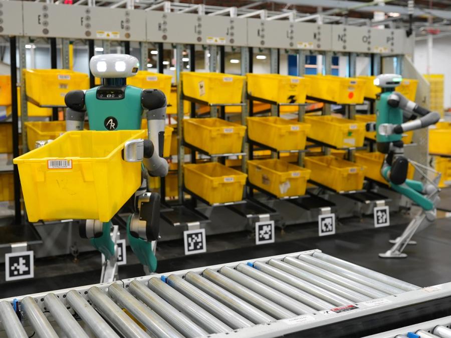 Two bipedal robots move yellow bins in a factory setting.