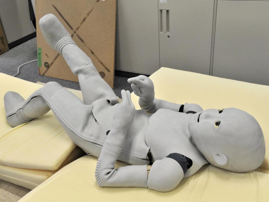 A grey fleshed toddler sized humanoid robot lies down on cushions.