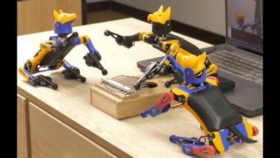 Three small robot dogs with yellow, black, and blue plastic bodies sit on a desk around a xylophone.