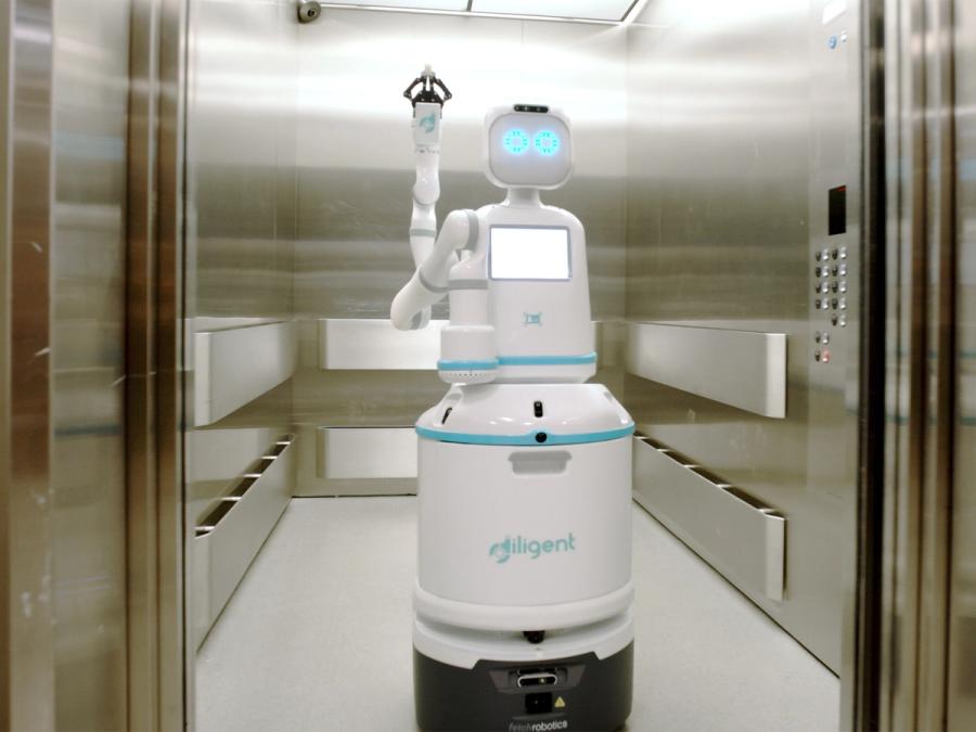 A white mobile robot with glowing circular shaped eyes and an articulated arm with a two finger gripper rides an elevator.