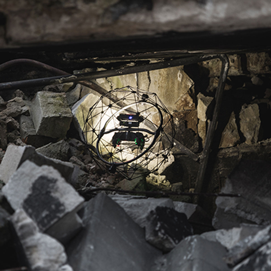A drone in a protective cage flies through building rubble.