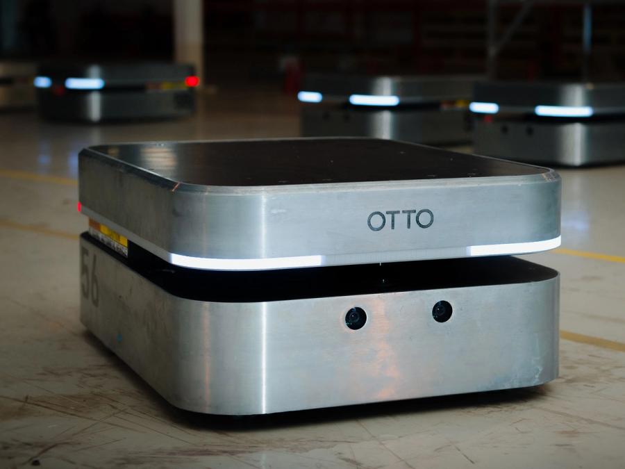 A squat, square, silver mobile robot with two layers, lights, and the name OTTO on it on a factory floor with many other behind it.