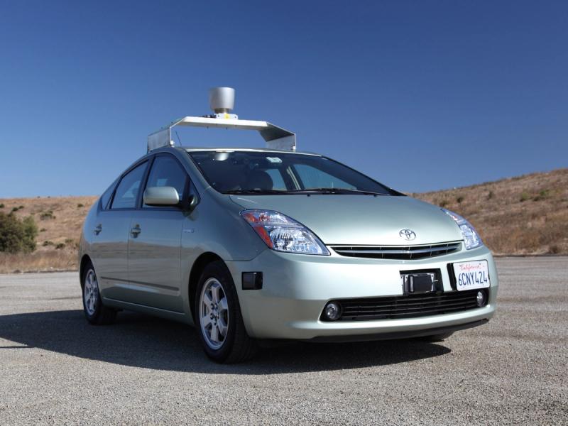 A car with cameras, radar, and a roof rack holding a large lidar instrument sits on a road.