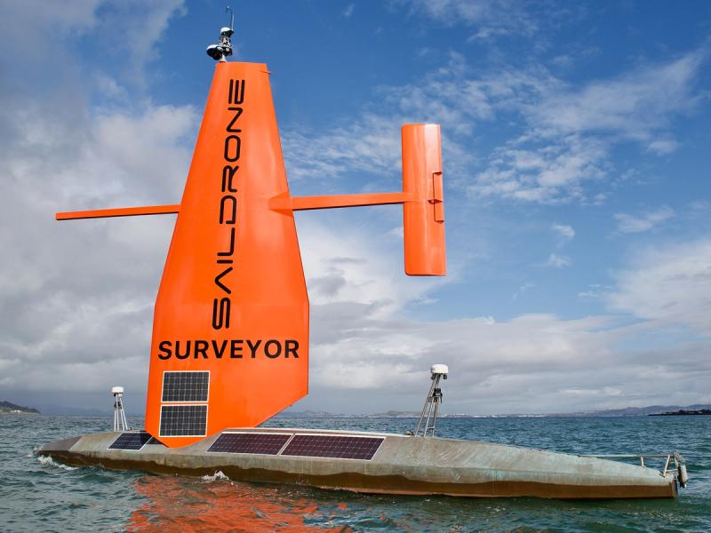 An uncrewed surface vehicle on the water. It has a horizontal silver unit with multiple solar panels and a large orange sail with solar panels rising vertically, and a rudder running parallel to the sea.