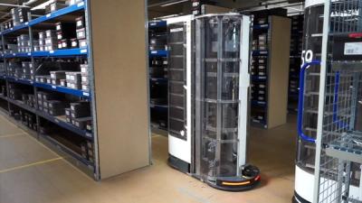 A tall mobile robot, with metal racks covered by a transparent plastic shell, drives in a narrow row between racks of shoes in a warehouse.