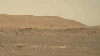 Ingenuity helicopter seen very small at the center of the frame flying over the rusty rocky surface of Mars.